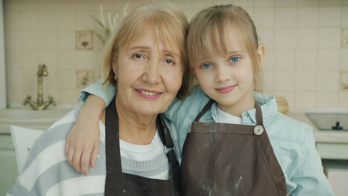 Portrait of grandmother and granddaughter in aprons smiling looking at camera in kitchen. Family lifestyle, happiness and happy childhood concept.